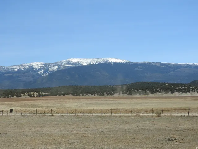 Expansive natural landscape featuring a rustic fence line, distant forested hills, and snow-capped mountains under a clear sky