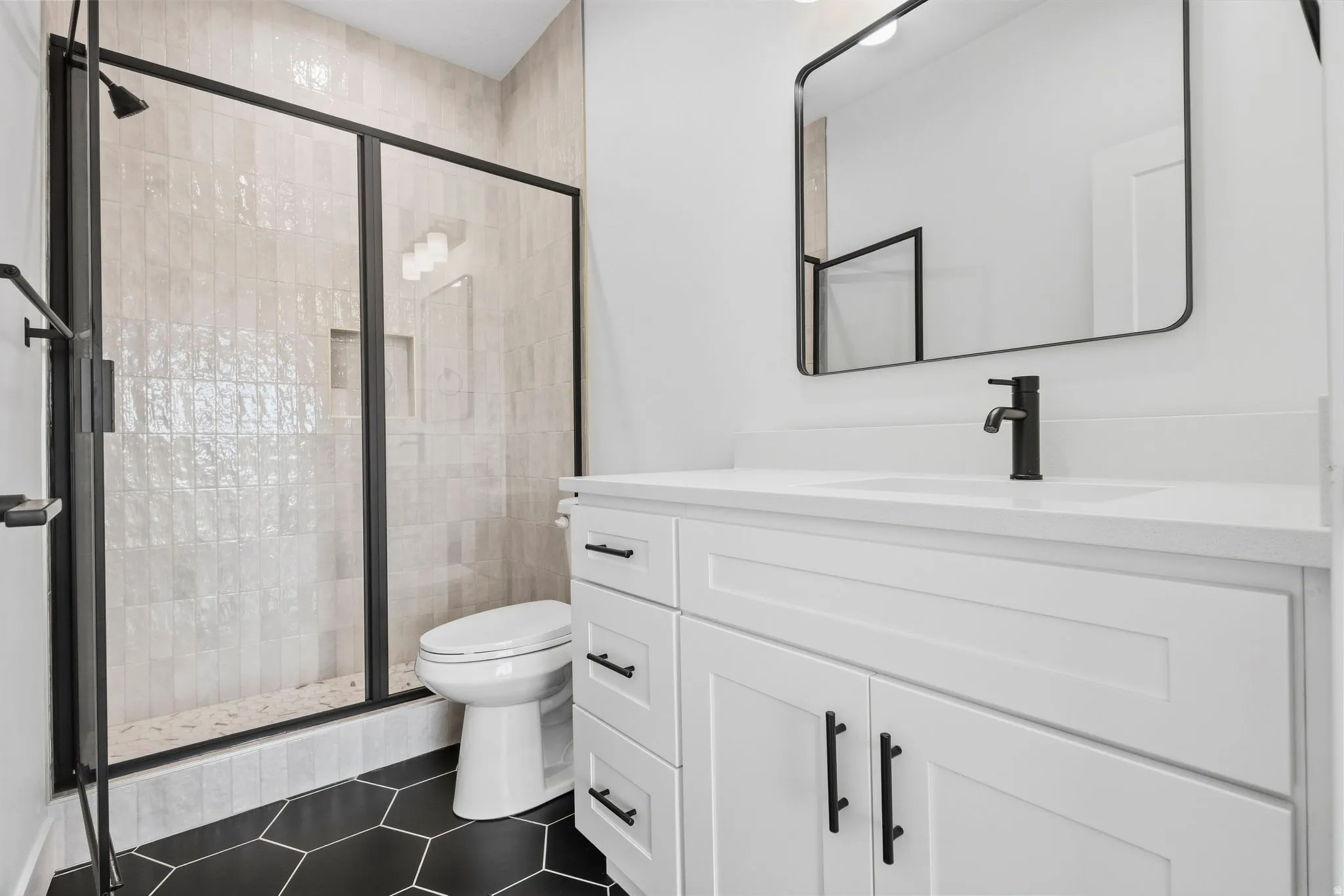 Matte black hexagonal floor tiles, white shaker-style vanity with integrated sink, sleek black framed mirror, and a glass-enclosed shower with vertical tiled walls