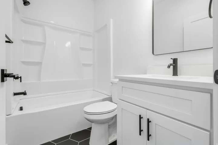 White vanity with a solid surface countertop, matte black fixtures, framed mirror, white tub, and hexagonal floor tiling
