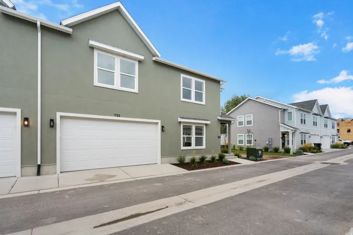 Stucco exterior, white frame windows, attached garage, exterior wall lighting, and manicured landscaping