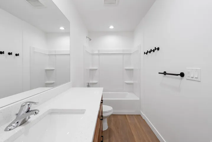 White bathroom featuring a built-in tub with white surround, wood-finish flooring, a vanity with a white countertop and integrated sink, and black hardware