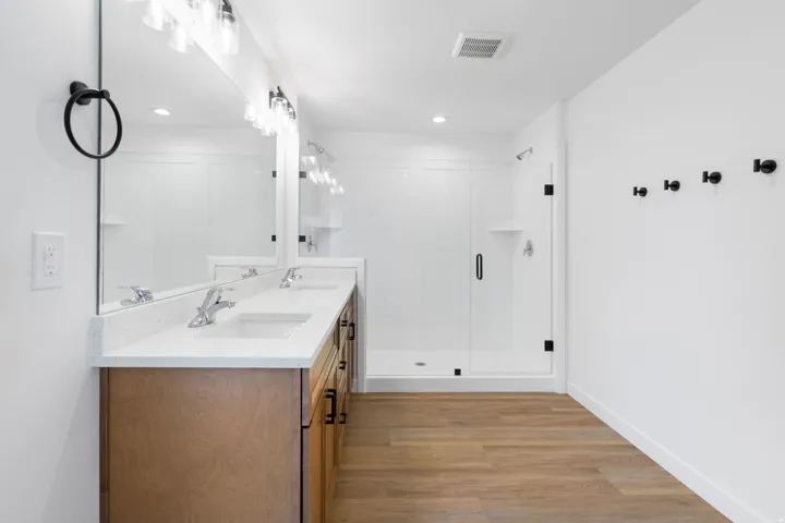 Dual vanity with integrated sinks and a light-toned countertop, wood-finish flooring, a glass-enclosed shower, and modern wall-mounted light fixtures
