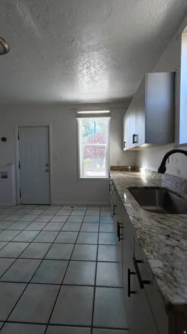 White cabinetry with black hardware, stainless steel sink, patterned countertop, tile flooring, and white door