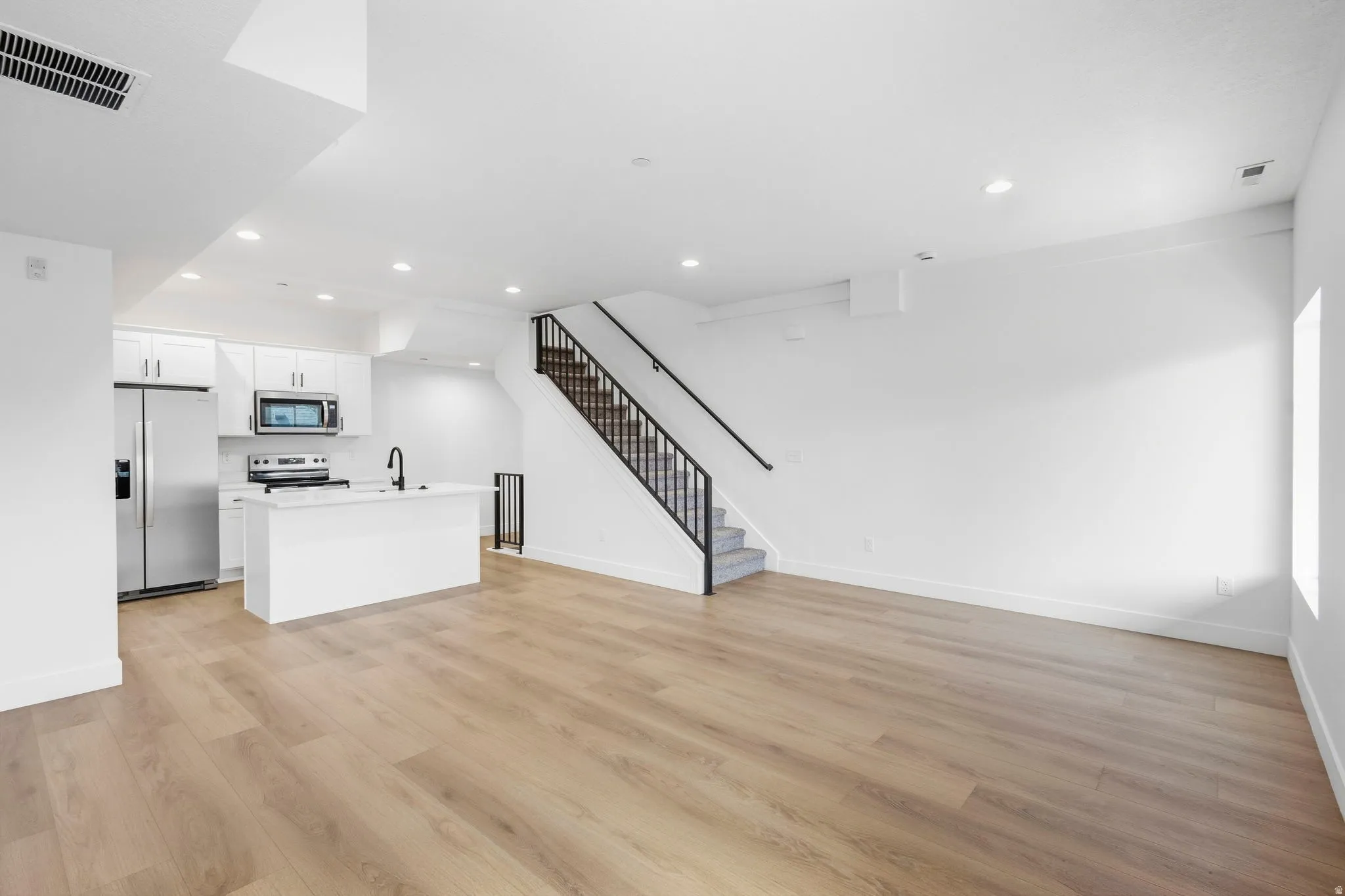 Open-concept living space with wood-finish flooring, white cabinetry, stainless steel appliances, and a staircase with a black metal railing