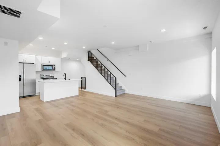 Open-concept living space with wood-finish flooring, white cabinetry, stainless steel appliances, and a staircase with a black metal railing