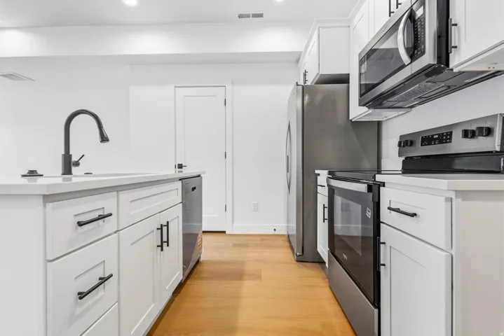 White cabinetry with matte black hardware, stainless steel appliances, light-toned wood-finish flooring, and a kitchen island with an integrated sink