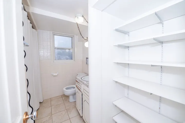 White adjustable shelving system, white toilet, white vanity with integrated sink, patterned wall tile, and a window