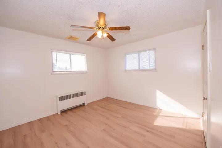 Light wood-finish flooring, white walls, two windows, ceiling fan with light, and a baseboard heating unit
