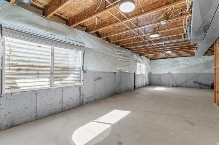 Unfinished concrete basement with exposed wood joists and installed lighting, featuring two windows with blinds