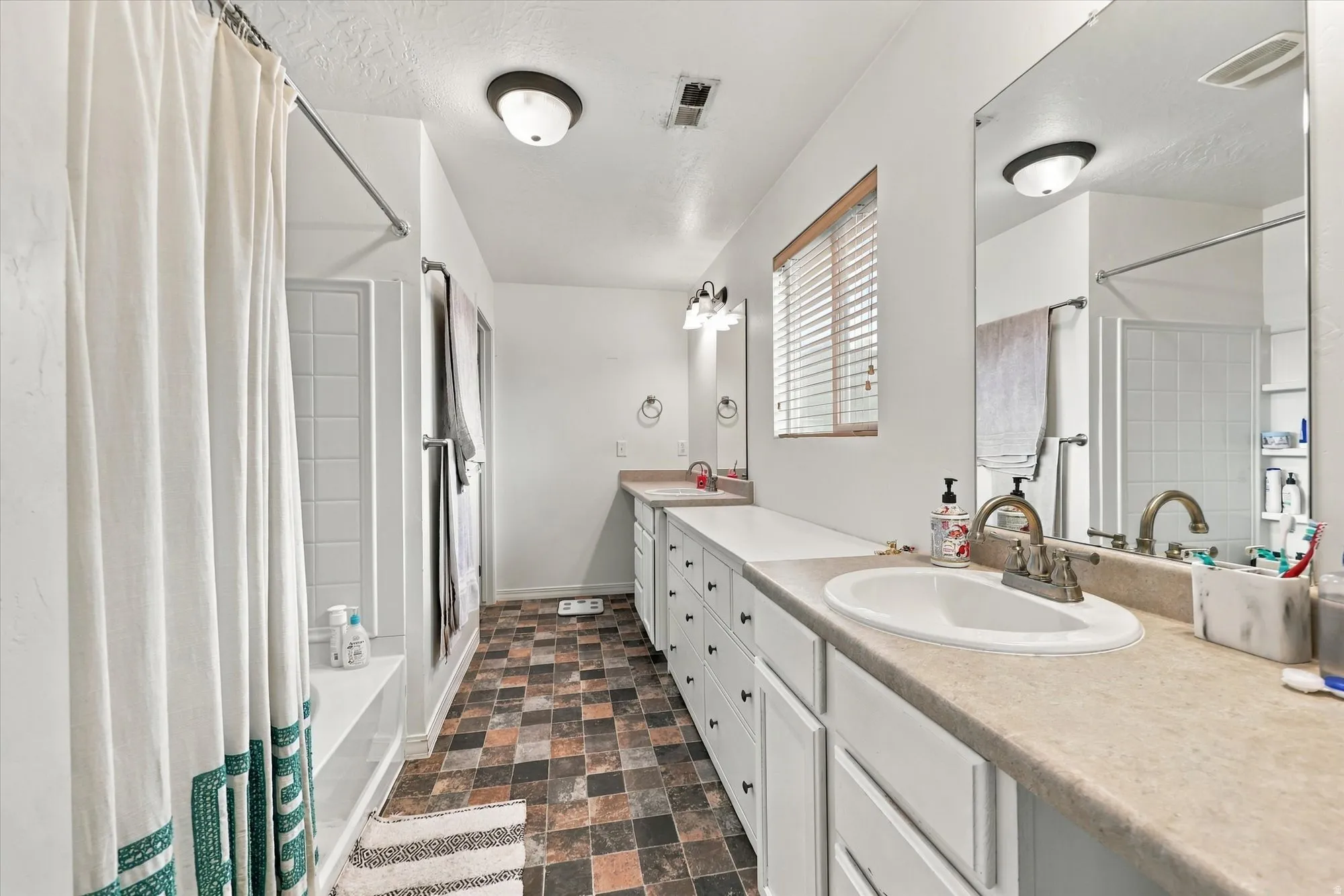 White paneled cabinetry with multiple drawers, dual integrated sinks with chrome fixtures, full-length mirror, and a window with horizontal blinds