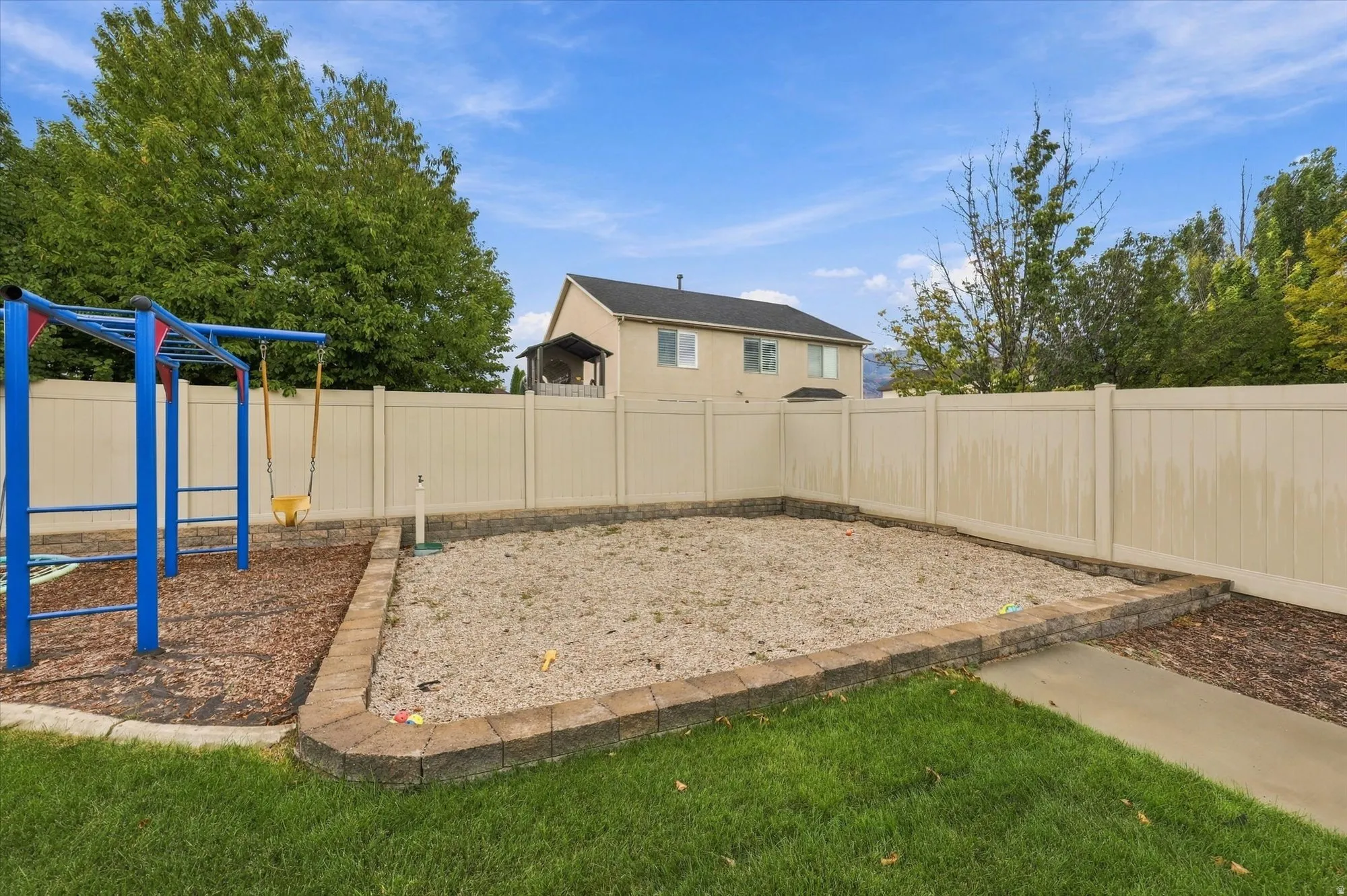 Fenced backyard featuring a gravel-filled area with decorative stone edging, a separate bark-mulch area with a blue play structure, and a concrete pathway