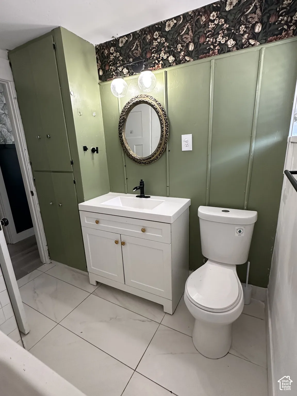 White tile flooring, white vanity with integrated sink, black finish faucet, ornate framed mirror, and built-in storage cabinetry