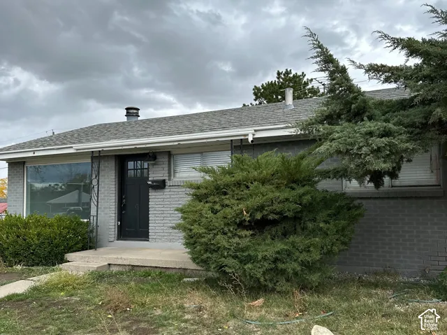 Single-story exterior featuring grey brick masonry, a covered concrete porch, a multi-lite front door, and a shingle roof