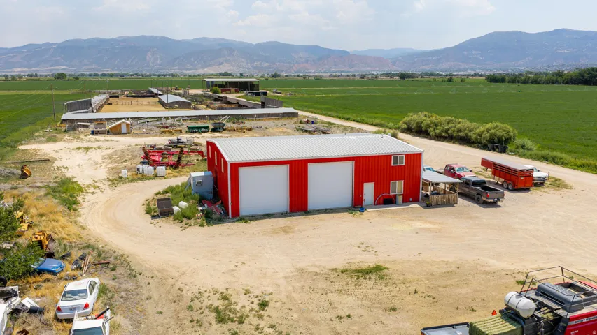 Red metal building with white garage doors, two-story structure, expansive dirt lot, surrounding green fields, and distant mountains