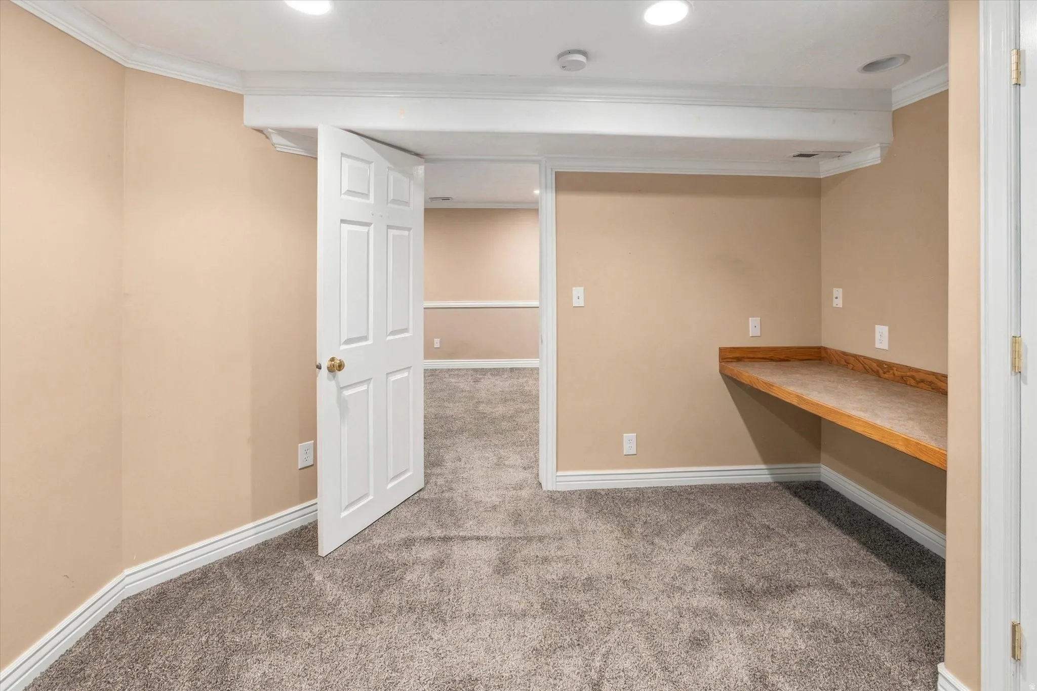 Recessed lighting illuminates the carpeted room, featuring crown molding and a built-in wood-finish desk