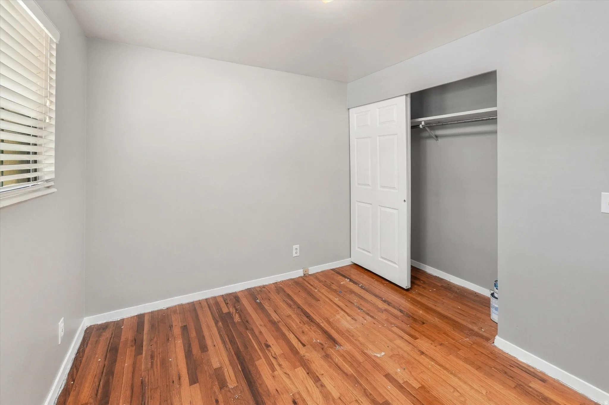 Hardwood flooring, light gray wall paint, window with horizontal blinds, and a built-in closet with sliding panel doors and an interior hanging rod