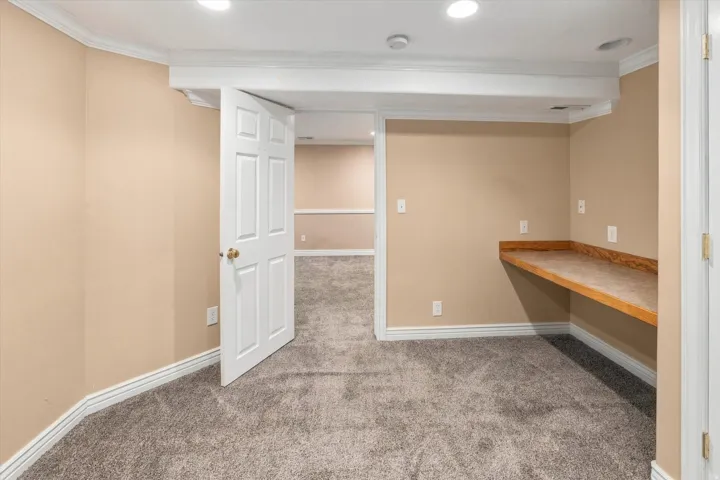 Recessed lighting illuminates the carpeted room, featuring crown molding and a built-in wood-finish desk