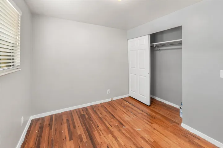 Hardwood flooring, light gray wall paint, window with horizontal blinds, and a built-in closet with sliding panel doors and an interior hanging rod
