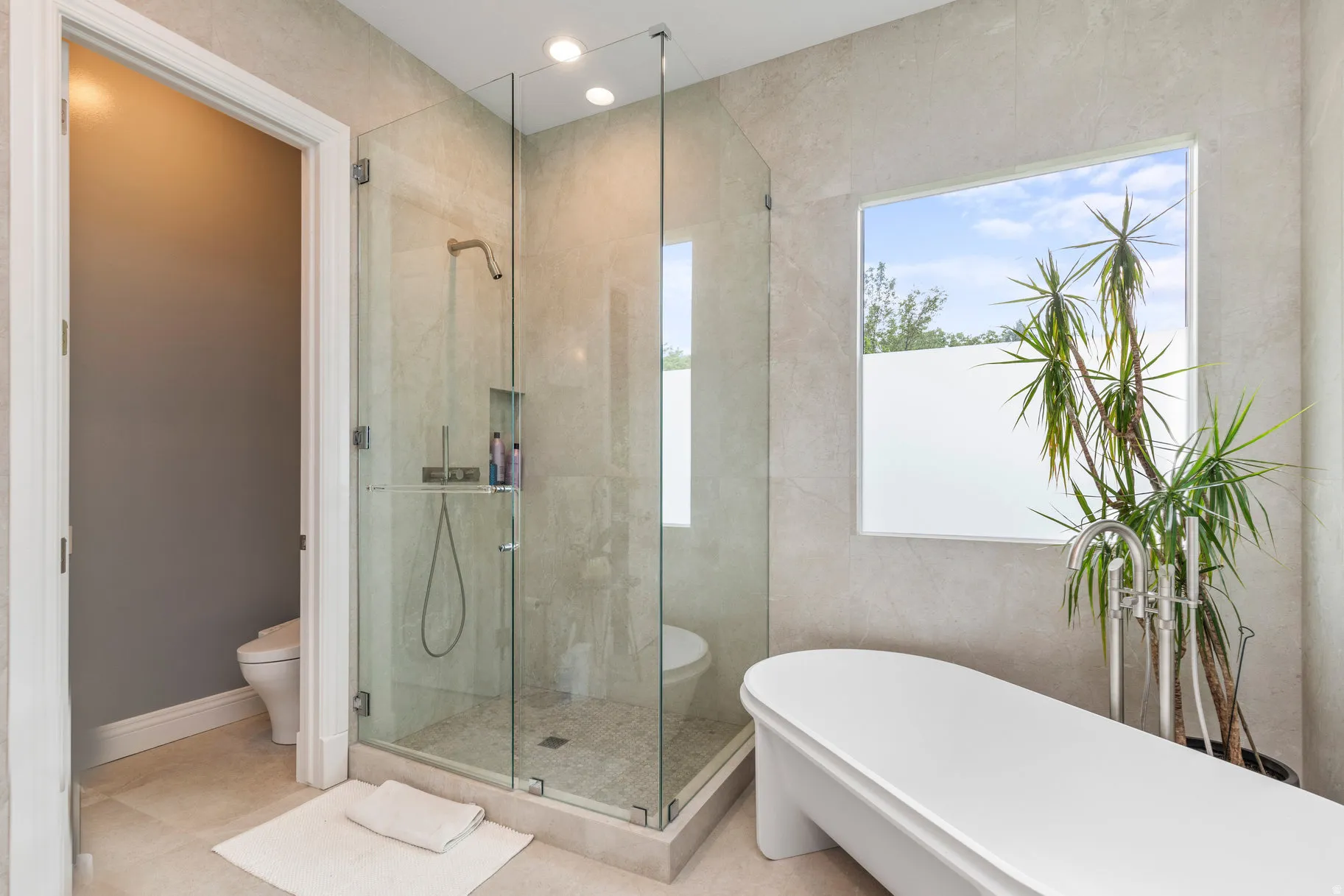 Freestanding soaking tub with floor-mounted faucet, expansive glass-enclosed shower, and a large window