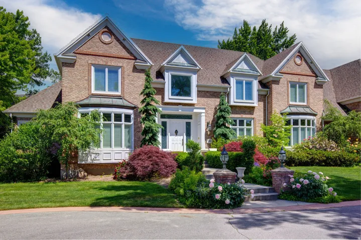 Brick exterior with multiple gables and bay windows, featuring a landscaped front yard with mature plantings and a paved driveway