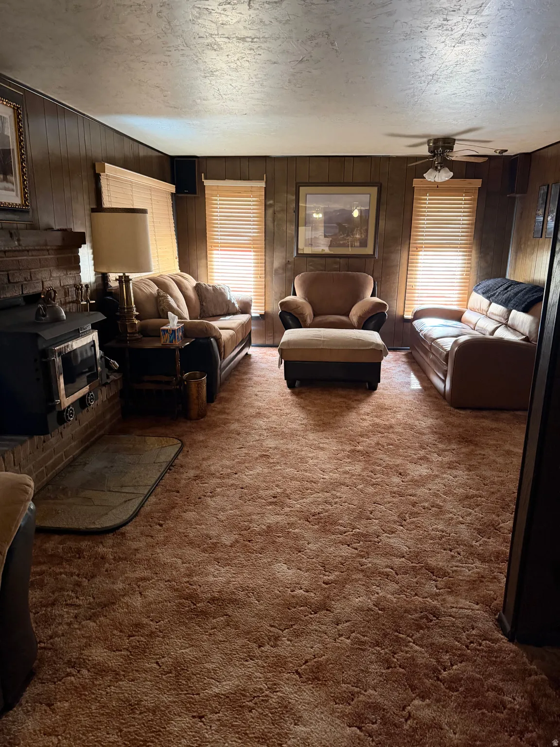 Wood-paneled walls, brick fireplace surround, built-in wood-burning stove, and ceiling fan with lighting fixture
