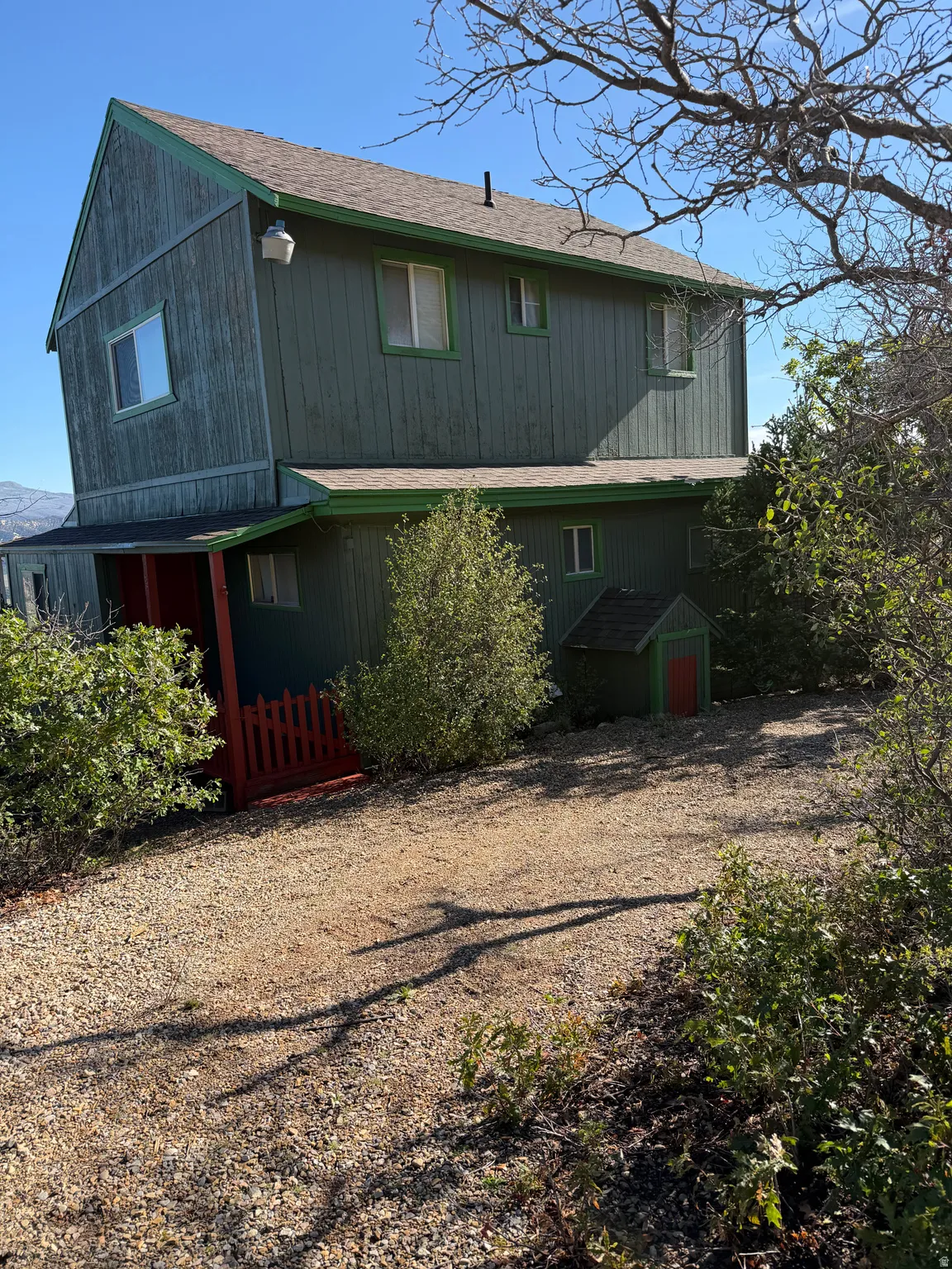 Multi-level exterior with green siding and white-framed windows, featuring a gravel driveway and mature trees