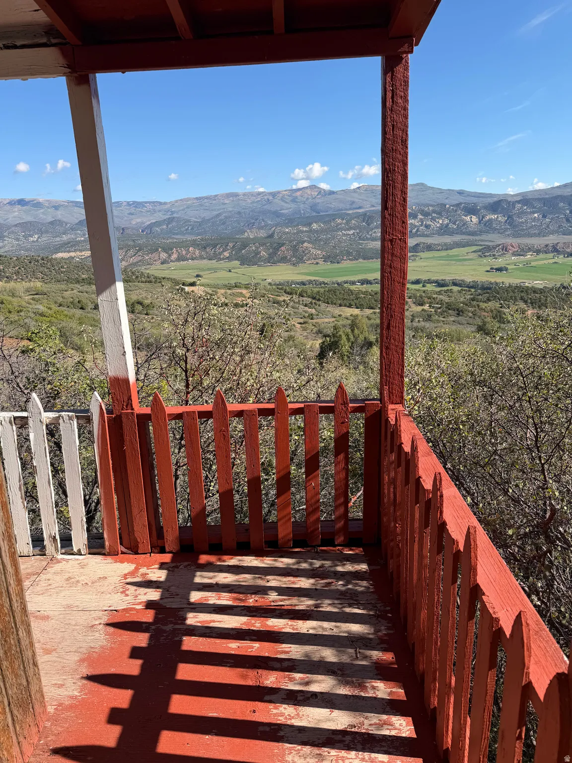 Balcony with a red picket fence railing and painted floor, framed by wood posts, offering expansive mountain and valley scenery