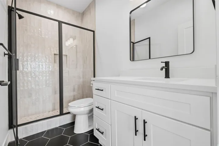 Matte black hexagonal floor tiles, white shaker-style vanity with integrated sink, sleek black framed mirror, and a glass-enclosed shower with vertical tiled walls