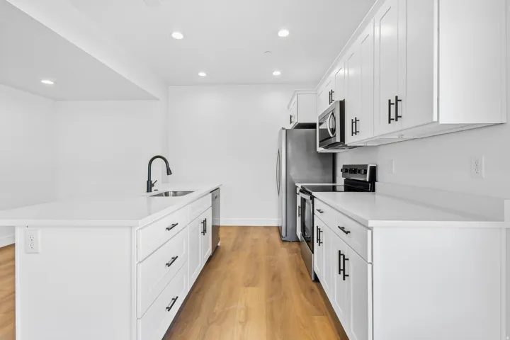 Kitchen featuring white cabinetry with black hardware, stainless steel appliances, a kitchen island with an integrated sink, and wood-finish flooring