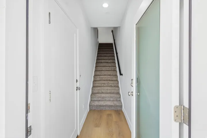 Light-toned wood-finish flooring, white walls, carpeted staircase, black metal handrail, and a frosted glass door
