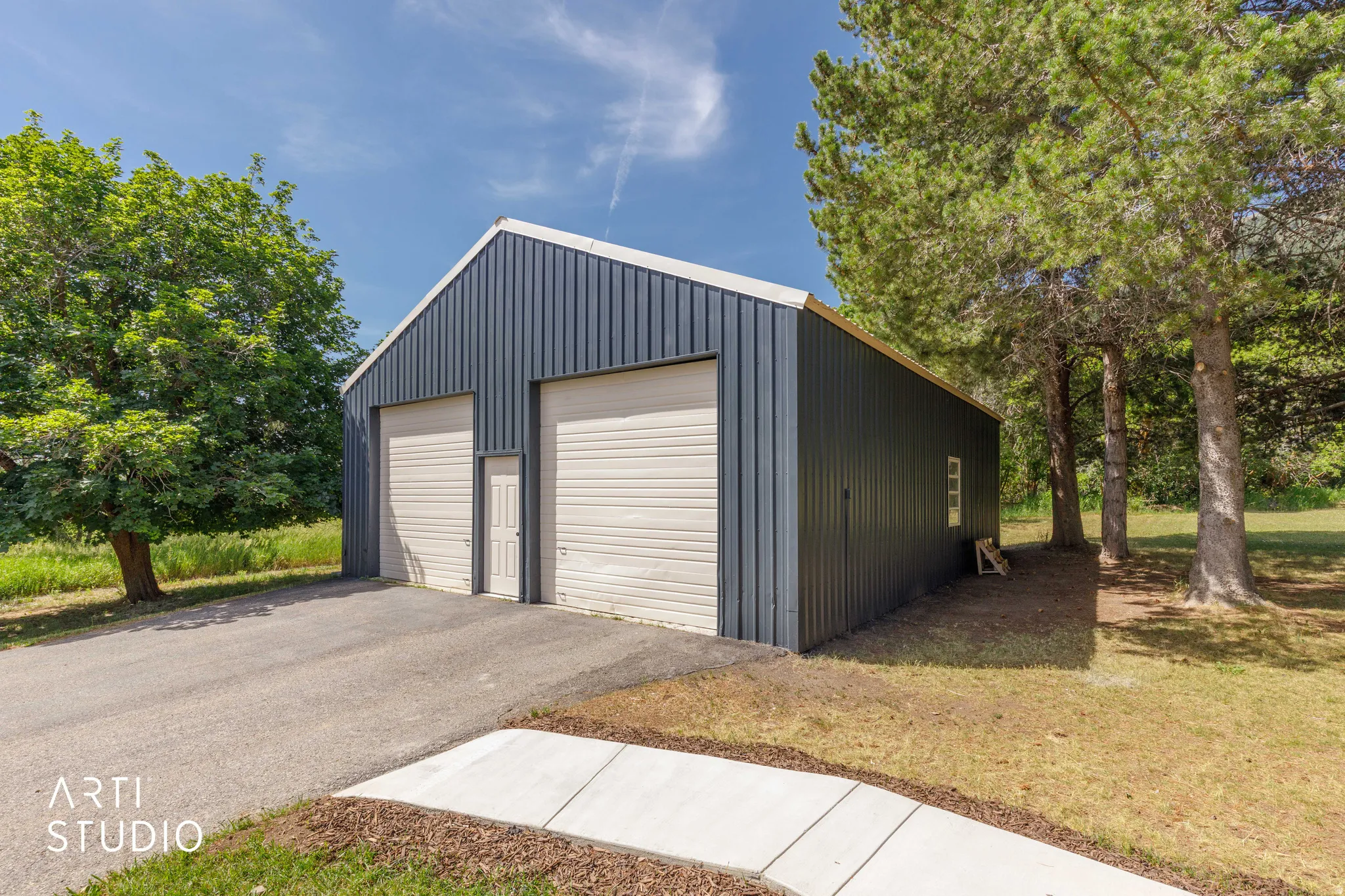Metal-sided outbuilding with two roll-up garage doors, a pedestrian entry door, and an asphalt driveway leading to a concrete pathway