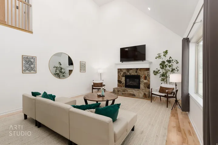 Vaulted ceiling, stone-faced fireplace with white mantle, wood-finish flooring, large window, and an interior balcony