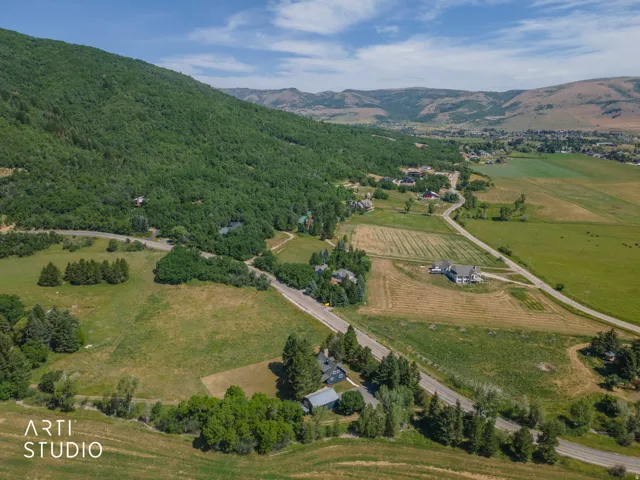 Expansive rural landscape with a winding paved road, surrounded by mature trees, vast green fields, and distant mountains