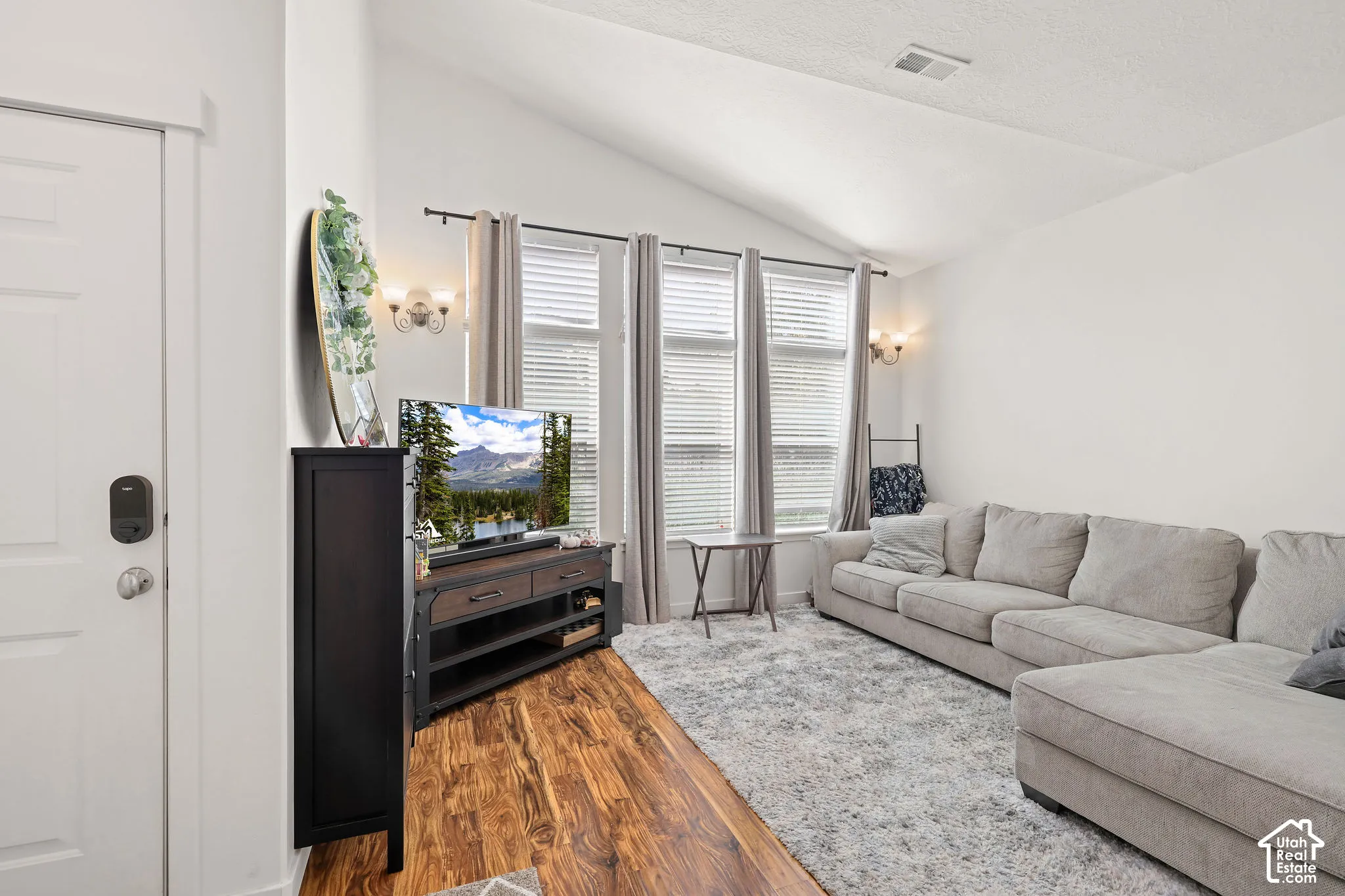Vaulted ceilings, wood-finish flooring, three vertical windows with blinds, and wall-mounted sconce lighting