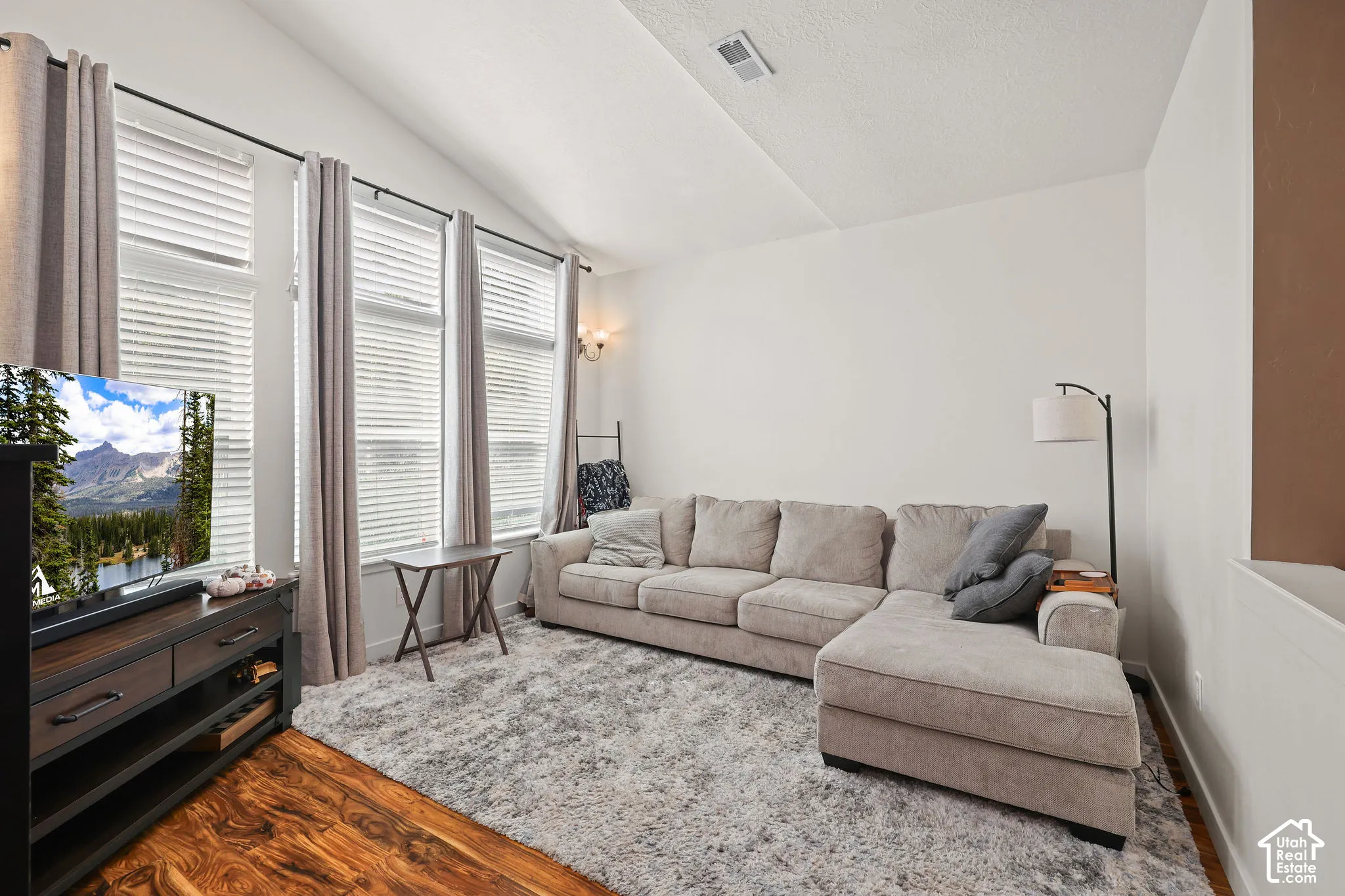Vaulted ceiling, wood-finish flooring, multiple windows with blinds, and white wall finishes