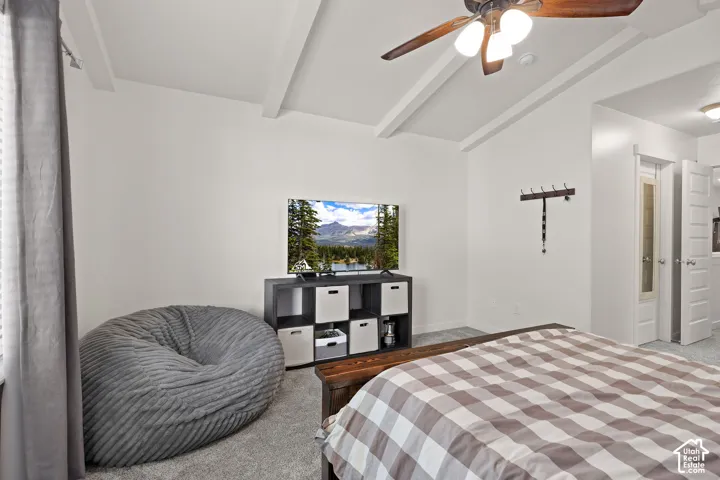 Vaulted ceiling with exposed beams, ceiling fan light fixture, and light gray carpeting