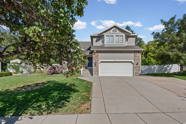 Stucco and stone facade, two-car garage, mature trees, manicured lawn, and concrete driveway