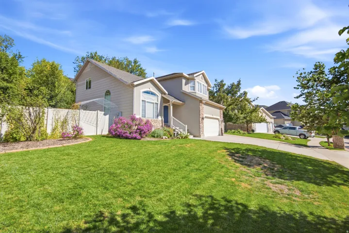 Two-story residential structure featuring a stucco and stone exterior, an attached garage, a gabled roofline, a front lawn, and foundational landscaping with flowering shrubs