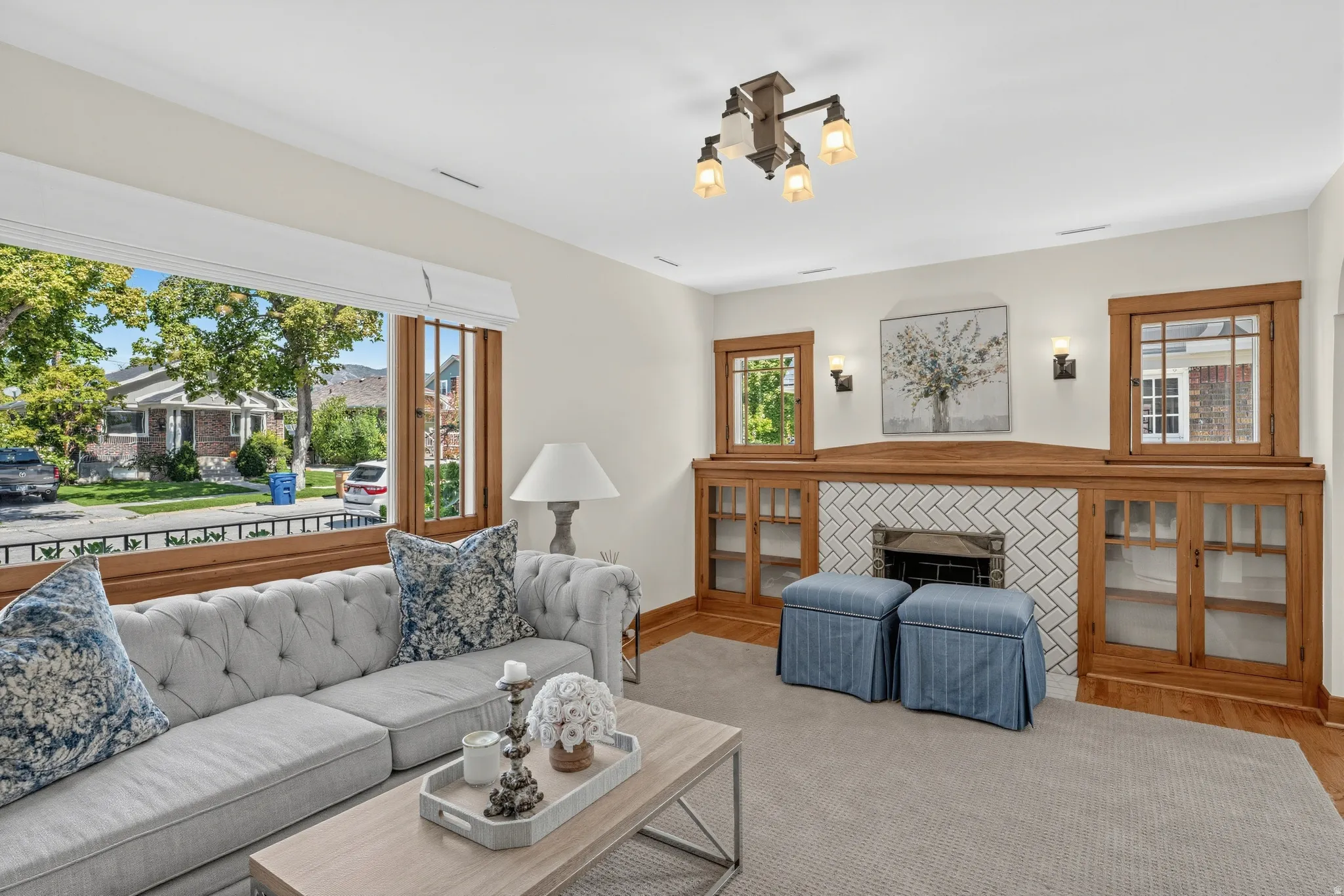 Expansive front-facing windows, built-in wooden cabinetry with glass inserts, herringbone tile fireplace surround, wood-finish flooring, and a ceiling-mounted light fixture