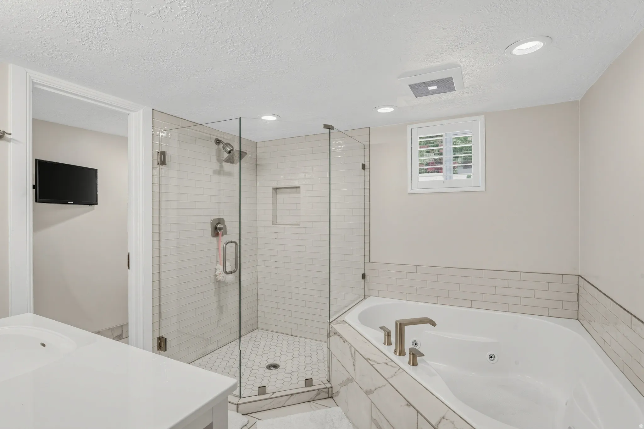 Walk-in shower with clear glass enclosure and subway tile surround, built-in corner bathtub with brushed nickel fixtures, single vanity with white countertop, recessed lighting, and a window with plantation shutters