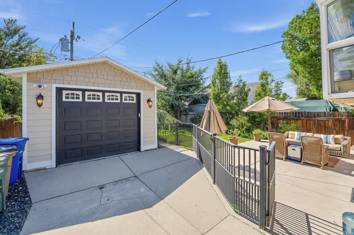 Detached garage with a dark panel door and decorative lighting, concrete driveway, concrete patio, and black metal fencing
