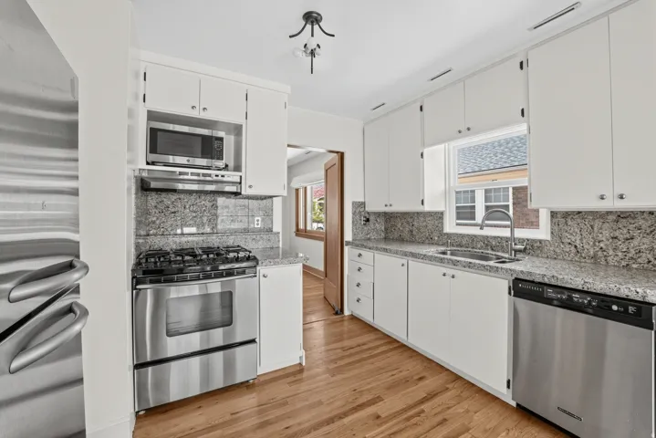 Stainless steel appliances, wood-finish flooring, white cabinetry, granite countertops, and a window above the sink