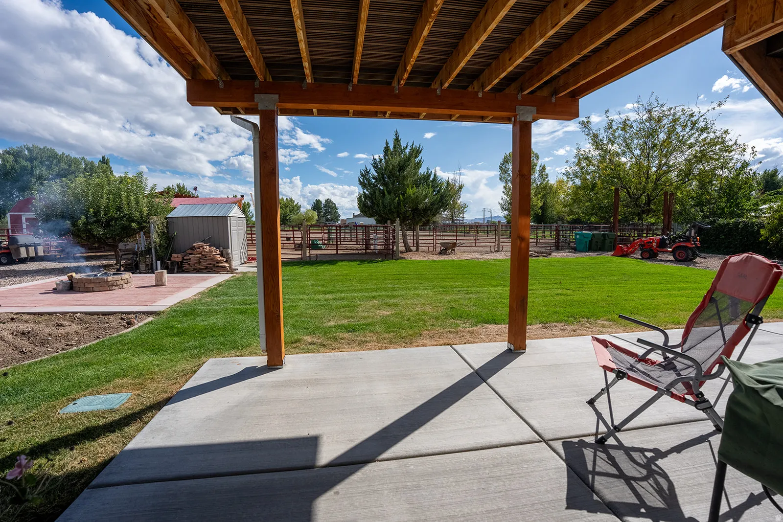 Covered patio with concrete flooring, overlooking a green lawn, brick paver patio with a built-in fire pit, and an outdoor storage shed