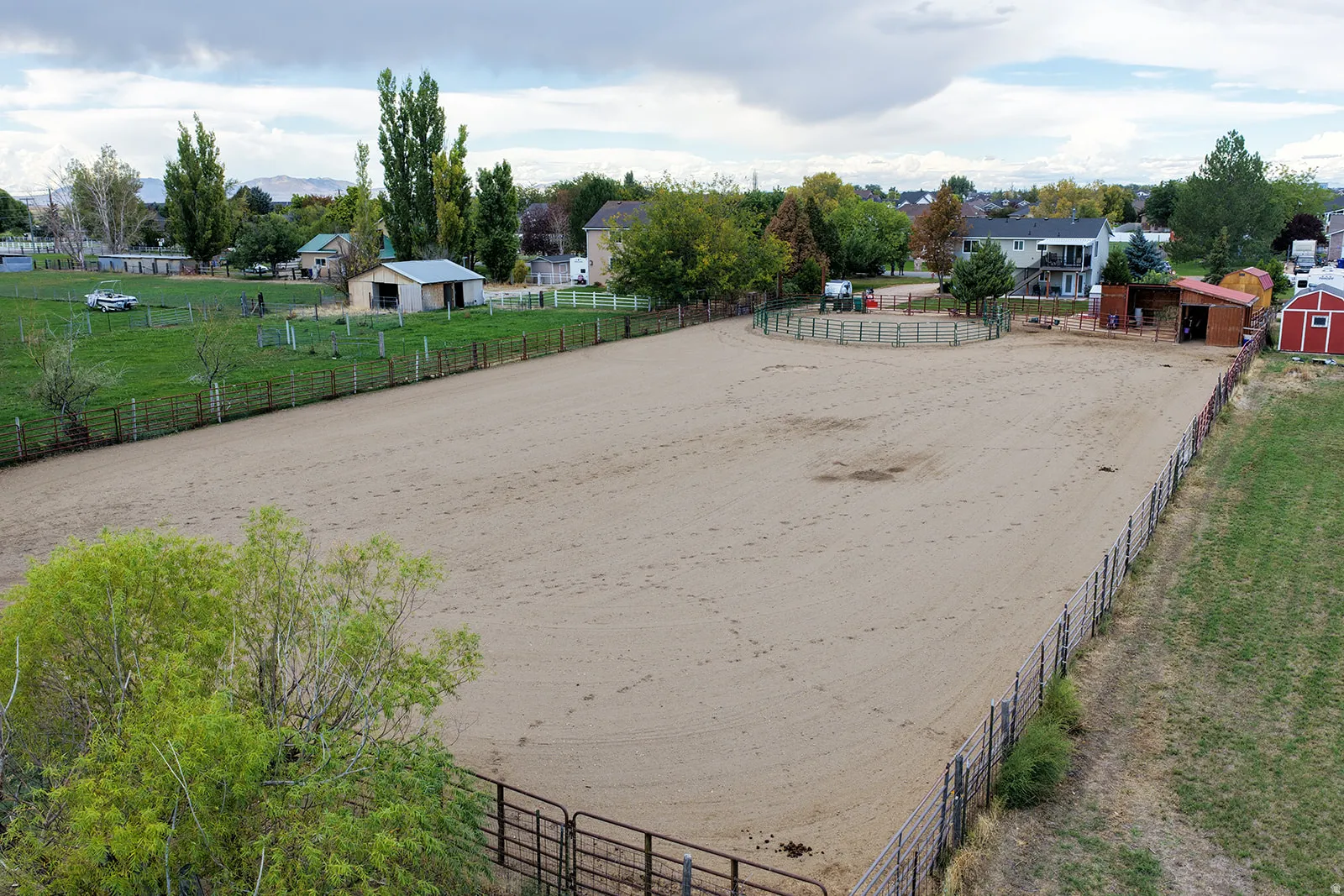 Expansive outdoor arena with sand footing, enclosed by fencing, featuring a round pen and a red barn structure