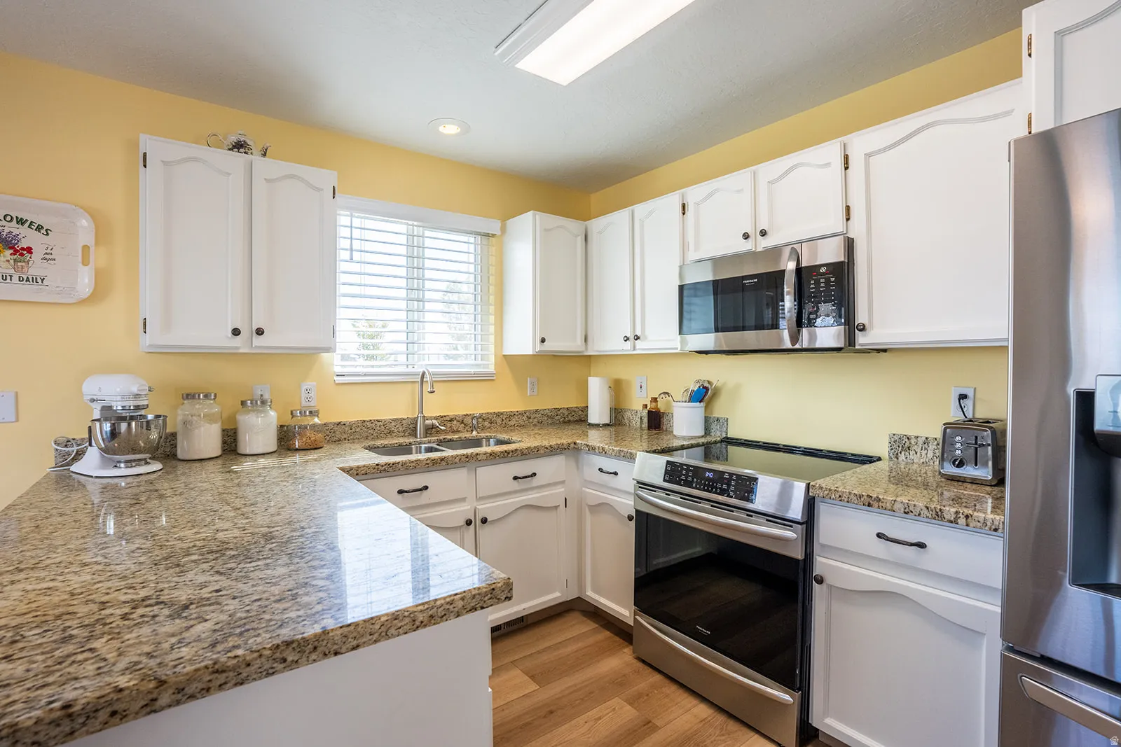 White cabinetry with dark hardware, stainless steel appliances, granite countertops, wood-finish flooring, and a double-basin sink