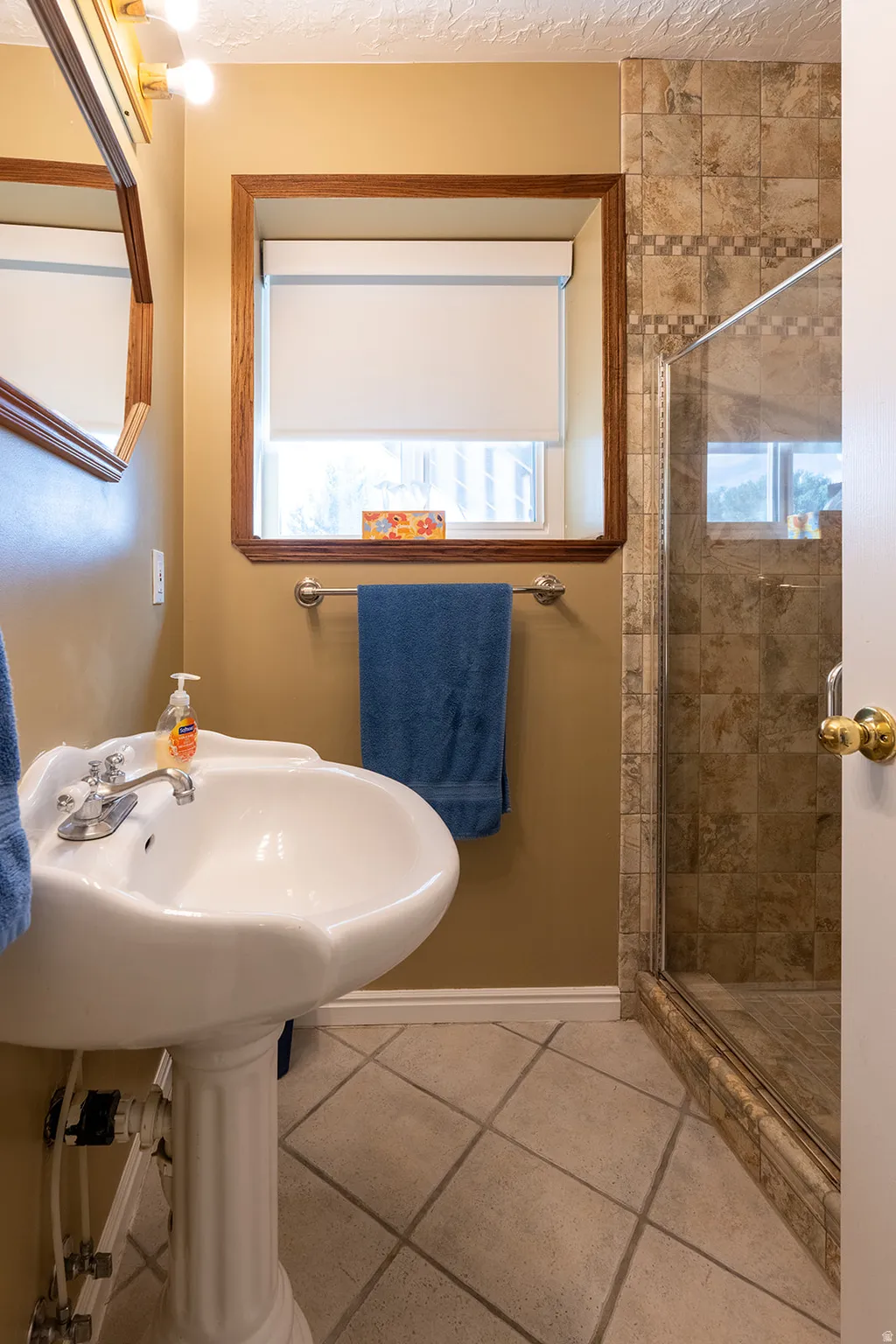 Pedestal sink, tiled flooring, glass-enclosed shower, wall-mounted light fixture, and a wood-framed mirror