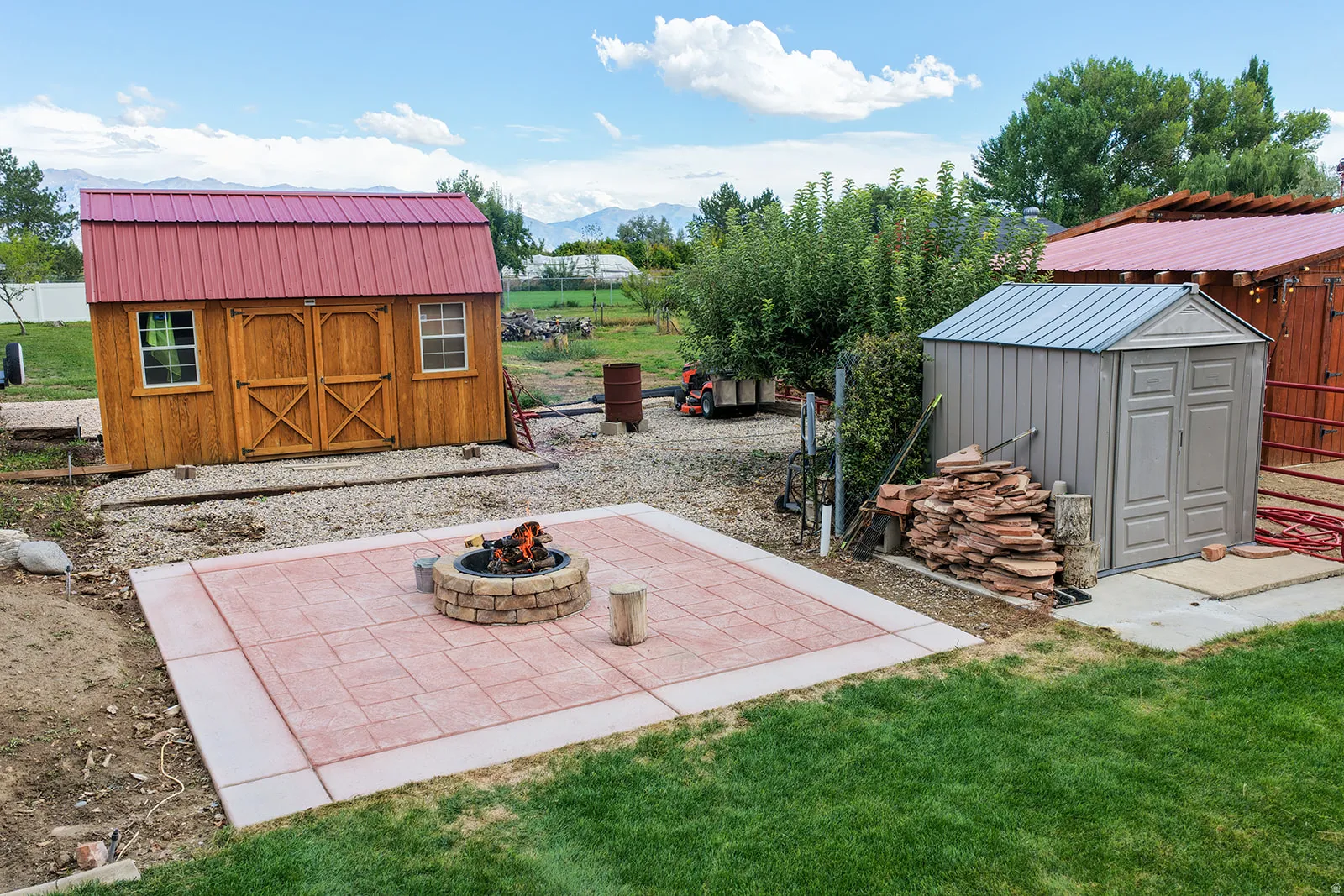 Expansive outdoor area featuring a concrete patio with a built-in fire pit, a wooden storage shed with a red metal roof, and an additional metal storage shed