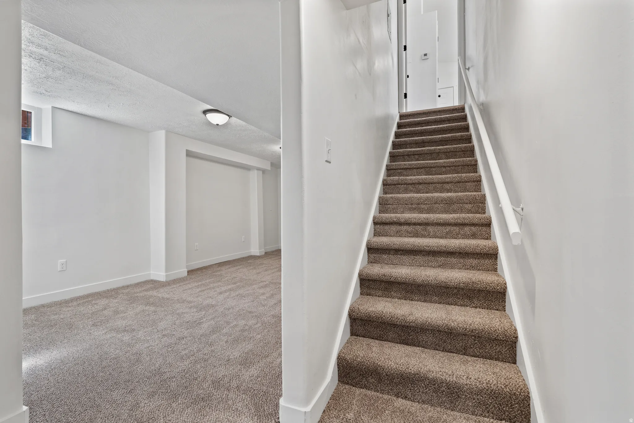 Carpeted stairs with a white handrail, leading to a carpeted room with white walls and a ceiling-mounted light fixture