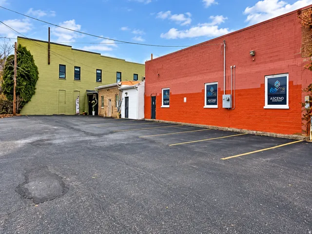 Paved parking area featuring striped parking spaces, with a multi-story building featuring green-painted brick and multiple windows, and an additional building with red-painted brick and window signage
