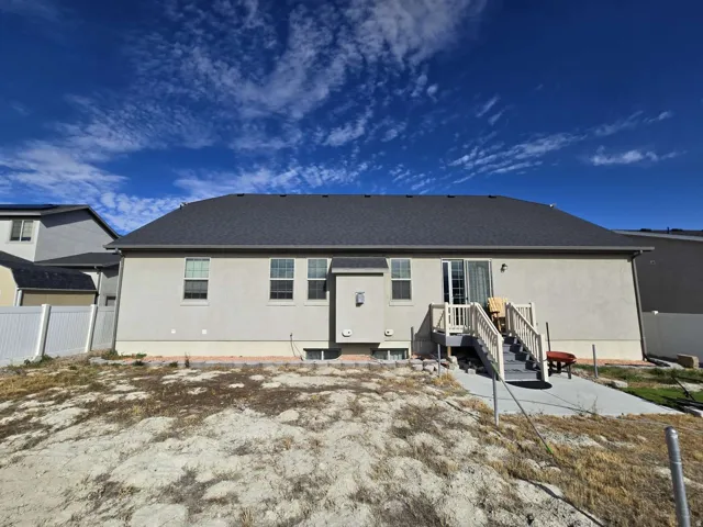 Exterior featuring a dark shingle roof, light-colored stucco siding, multiple windows, a rear deck with stairs, and a concrete patio